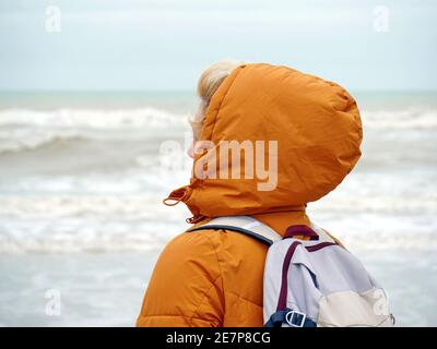 Eine junge Frau mit einem Rucksack in einer gelben Jacke und einer Kapuze auf dem Kopf blickt auf die Wellen des Ozeans. Wolkenloses Wetter in Dieppe, Frankreich Stockfoto