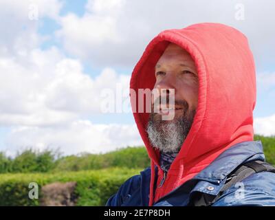 Ein bärtiger Mann in einer roten Kapuze schaut nachdenklich hinein Die Distanz und lächelt Stockfoto