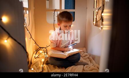 Netter kleiner Junge liest Buch mit Fackel in seinem Spielzeug Papphaus in der Nacht. Konzept der Kindererziehung und Lesen im dunklen Raum Stockfoto