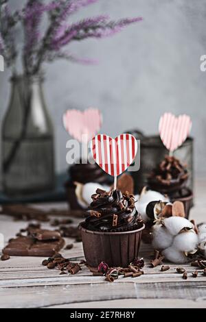 Mehrere Muffins oder Cupcakes mit schokoladenförmiger Sahne am weißen Tisch. Eine Karte in Form eines Herzens zum Valentinstag. Die Hand einer Frau zerbröckelt Stockfoto