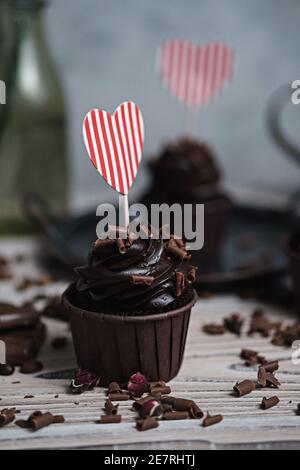 Mehrere Muffins oder Cupcakes mit schokoladenförmiger Sahne am weißen Tisch. Eine Karte in Form eines Herzens zum Valentinstag. Die Hand einer Frau zerbröckelt Stockfoto