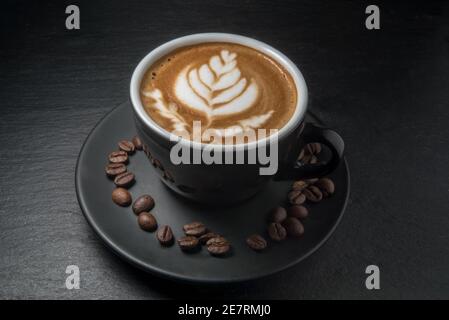 Kaffee in schwarzer Tasse mit Blattbild auf Schaum und mit Kaffeebohnen auf Untertasse, schwarzer Schieferstein Hintergrund Stockfoto