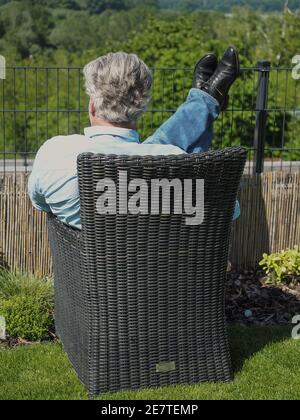 Leitender kaukasischer Geschäftsmann mit grauen Haaren sitzt auf einem Stuhl im Garten. Sonniger Frühlingstag. Vertikale Aufnahme. Stockfoto