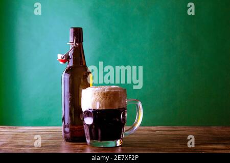 Ein Becher dunkles Bier mit seiner offenen Flasche auf grünem Hintergrund und einem Holztisch. St. Patrick und Stout Konzept Stockfoto