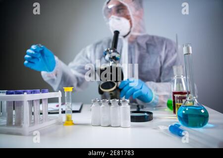 Wissenschaftler oder Sanitäter in Schutzkleidung, nimmt Reagenzglas mit blauem Reagenz, Laborglas mit chemischer Flüssigkeit, Mikroskop, Labor Stockfoto