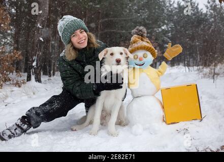 Happy cute Teenager Mädchen, weißer Hund, fröhlich Schneemann in gelb gestrickt Hut, Jacke, Fäustlinge, medizinische Maske mit Lächeln auf sie in einem verschneiten Wald gemalt. Stockfoto