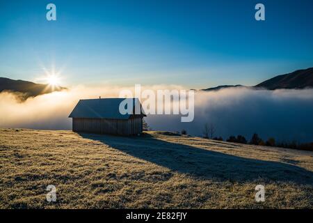 Ein einsames kleines Haus steht auf einer gefrorenen Wiese vor dem Hintergrund eines wunderschönen blauen Himmels, heller Sonne und weißem Nebel Stockfoto