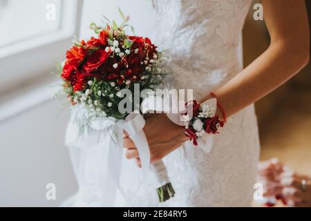 Schuss einer Braut Hand mit boutonniere hält eine schöne Rotes Bouquet mit weißem Band Stockfoto