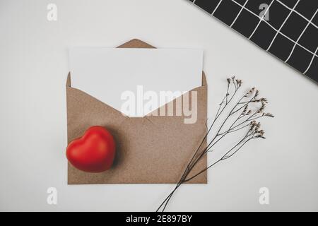 Weißes Blankopapier wird auf einen offenen braunen Papierumschlag mit rotem Herz und Limonium Trockenblume und schwarzem Tuch auf weißem Hintergrund gelegt. Valentinstag Stockfoto