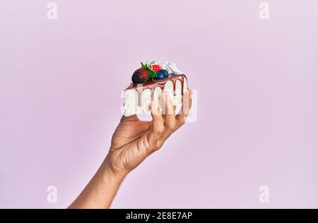 Hand des hispanischen Mannes hält Scheibe Schokoladenkuchen über isolierten rosa Hintergrund. Stockfoto
