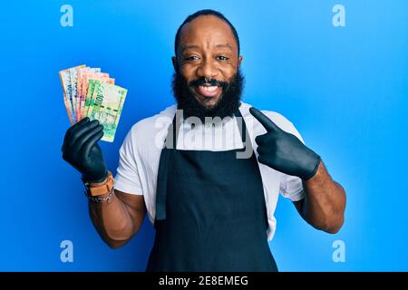 Junger afroamerikanischer Mann mit professioneller Schürze, der Südafrika hält rand Geldscheine lächelnd glücklich zeigen mit Hand und Finger Stockfoto