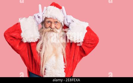 Alter älterer Mann mit grauen Haaren und langem Bart trägt traditionelle weihnachtsmann Kostüm posiert lustig und verrückt mit Fingern auf dem Kopf als Hase Ohren, Smilie Stockfoto