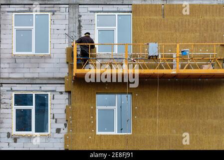 Ein Bauherr, der auf einer hängenden Plattform steht, dämmt die Fassade eines Hochhauses im Bau mit Mineraldämmung. Stockfoto