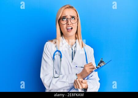 Schöne kaukasische blonde Frau trägt Arzt Stethoskop hält Zwischenablage wütend und verrückt schreien frustriert und wütend, schreien mit Wut Blick Stockfoto