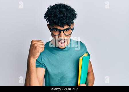 Junger araber schöner Mann trägt Brille und hält Bücher schreien stolz, feiert Sieg und Erfolg sehr aufgeregt mit erhobenen Armen Stockfoto