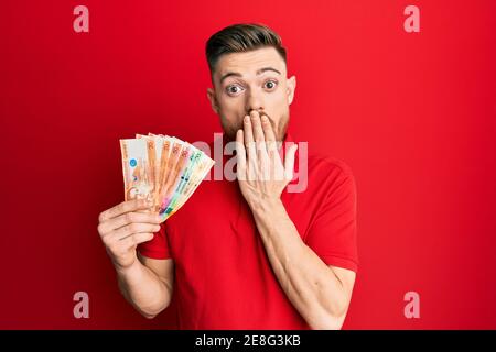 Junger Rotschopf Mann hält philippinischen Peso Banknoten über den Mund mit der Hand, schockiert und Angst vor Fehler. Überrascht Ausdruck Stockfoto