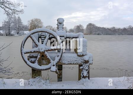 Verschneite historische Schleuse Rad auf einem gefrorenen gestauten Mühlenteich im Winter unter einem grau bewölkten Himmel, Kopierraum, ausgewählter Fokus Stockfoto