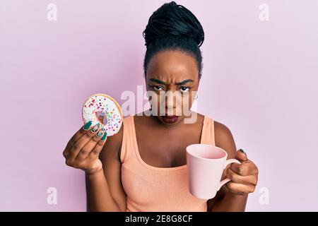 Junge afroamerikanische Frau essen Donut und trinken Kaffee ahnungslos und verwirrt Ausdruck. Zweifel Konzept. Stockfoto