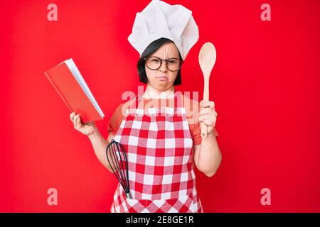 Brunette Frau mit Down-Syndrom tragen professionelle Bäcker Schürze Lesen Kochen Rezept Buch skeptisch und nervös, runzelte wegen des Problems. Stockfoto