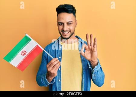 Junger arabischer Mann hält iran Flagge tut ok Zeichen mit Fingern, lächelnd freundlich gestikulieren ausgezeichnetes Symbol Stockfoto