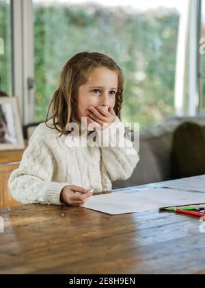 Cute erstaunt kleines Mädchen in weißen Pullover Abdeckung Mund mit Hand und wegschauen, während man am Tisch sitzt und zeichnet Mit Bleistiften Stockfoto