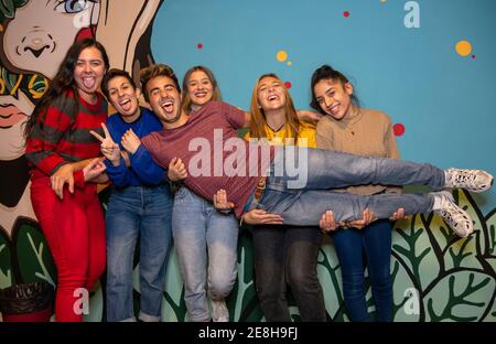 Gruppe aufgeregt junge Freunde halten Mann mit Zunge aus Auf Händen, während sie zusammen gegen eine helle Wand stehen Stockfoto