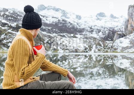 Rückansicht der anonymen ruhigen jungen Frau in warmen Pullover Und hat das Trinken von heißem Getränk, während Sie auf scharfem Stein sitzen Am kalten Seeufer umgeben Stockfoto