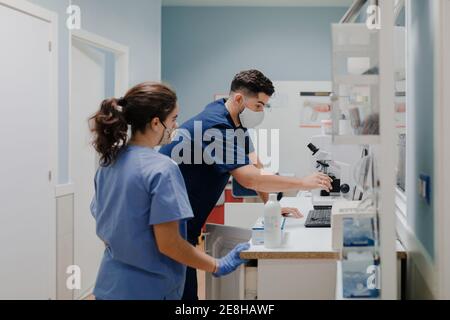 Seitenansicht des anonymen männlichen medizinischen Kropf in Uniform und Maske unter Verwendung des Mikroskops während der Arbeit im Labor in der Nähe einer Kollegin Stockfoto