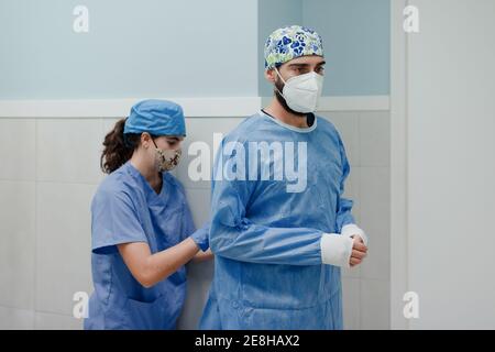 Unkenntlich weibliche Krankenschwester binden sterile Uniform auf männlichen Sanitäter in Atemschutzmaske vor der Operation im Krankenhaus Stockfoto