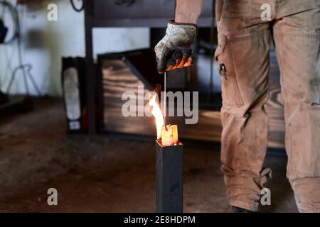 Crop anonymous tätowiert männlichen Schmied in lässiger Kleidung und Schürze Metallzangen während des Schmiedeprozesses in der Werkstatt in Flammen erhitzen Stockfoto