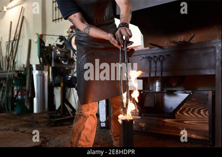 Crop anonymous tätowiert männlichen Schmied in lässiger Kleidung und Schürze Metallzangen während des Schmiedeprozesses in der Werkstatt in Flammen erhitzen Stockfoto