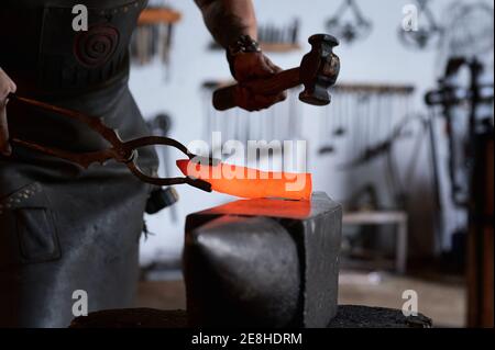 Seitenansicht des anonymen tätowierten jungen männlichen Fälschers in Schürze Erhitzen von Metall im Ofen während der Arbeit in der Grubbe Stockfoto