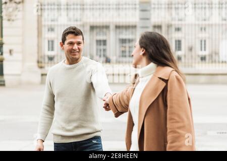 Fröhliches junges Paar, das warme Kleidung trägt und beim Gehen die Hände hält Gemeinsam auf Asphalt Bürgersteig in der modernen Stadt Stockfoto