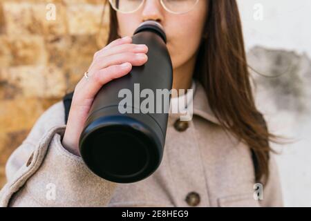 Crop junge Frau trägt warmen Mantel Trinkwasser aus schwarz Wiederverwendbare Flasche, während Sie am Herbsttag auf der Straße stehen Stockfoto