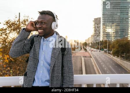 Junge fröhliche afroamerikanische männliche Büroangestellte in formeller Kleidung Hören Sie Musik mit Kopfhörern, während Sie in die Stadt blicken Stockfoto