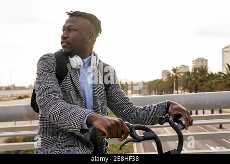 Junger nachdenklicher afroamerikanischer Manager mit Kopfhörern und Afro Haarschnitt mit dem Fahrrad in der Stadt Stockfoto