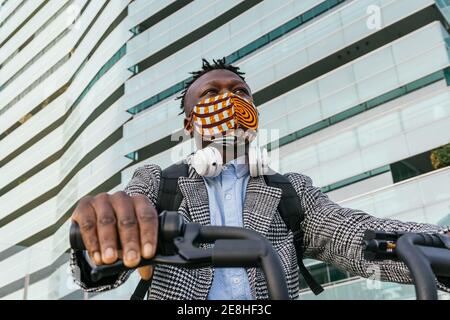 Niedriger Winkel der anonymen ethnischen männlichen Manager in Maske und Formell tragen Fahrrad Reiten gegen städtische Gebäude beim Blick weg Stockfoto