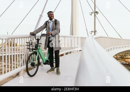 Junge lächelnde ethnische männliche Büroangestellte in formelle Outfit mit Kopfhörer und Fahrrad mit Blick auf die Hängebrücke in der Stadt Stockfoto
