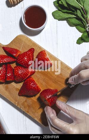 Draufsicht Crop unkenntlich Koch in Latex-Handschuhe Schneiden frisch Leckere Erdbeeren auf Holzschneidebrett auf dem Tisch Stockfoto