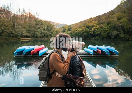 Seitenansicht junge liebevolle Paar in warmen tragen küssen zärtlich Während auf See hölzernen Kai gegen üppig reichlich bewaldet stehen Hügel im Stausee Valde Stockfoto