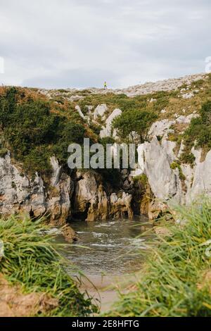 Malerischer Blick auf die Entfernung unkenntlich Person auf der Spitze stehen Berghang in der Nähe von kleinen sandigen Ufer des plätschernden schäumenden Meerwassers Bedeckt mit lu Stockfoto