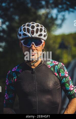 Selbstbewusster junger Radsportler in Sportbekleidung und Schutzhelm und Sonnenbrillen stehen grüne Bäume nach dem Training im Park auf sonnig Tag Stockfoto