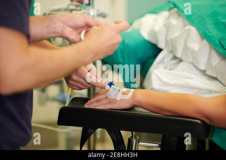 Schneiden Sie nicht erkennbare medizinische Assistenten, die intravenösen Katheter in der Hand auf Anonymer Patient, der im Operationssaal auf der Couch liegt Stockfoto