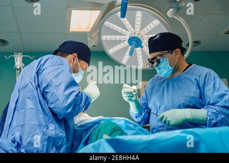 Nicht erkennbarer Patient, der während der Operation auf dem Operationstisch liegt Konzentrierte männliche Ärzte in medizinischen Kleidern und Masken Stockfoto