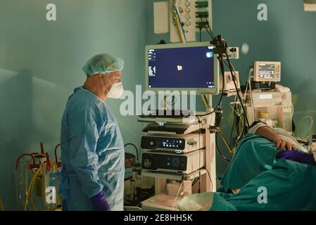 Seitenansicht von unkenntlich älteren männlichen Arzt in OP-Kittel Und Maske, die in der Nähe des Patienten auf dem Behandlungstisch vor der endoskopischen Verfahren Stockfoto