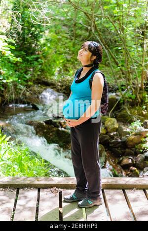 Seitenansicht der lächelnden schwangeren Touristen, die auf Holz stehen Brücke im Wald und genießen Sie die Natur während des Sommerurlaubs auf Hintergrund des schnellen Flusses Stockfoto