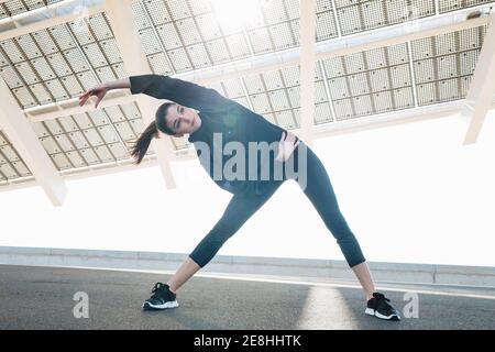 Positive fit junge Frau in schwarz Sportswear Stretching Arme während Wir wärmen uns auf einer sonnigen Straße in einem Vorort auf und schauen weg Zufrieden Stockfoto