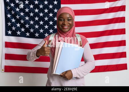 Lächelnde Frau in Hijab zeigt Daumen nach oben über amerikanische Flagge Stockfoto