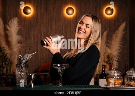 Fröhliche junge Barkeeper mit langen blonden Haaren in stilvoll In der modernen Bar werden in einem Shaker Cocktails gemischt und die Kleidung lächelt Stockfoto