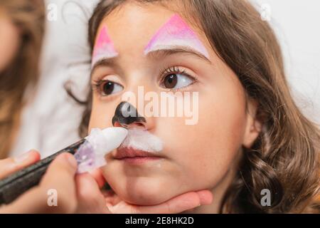 Crop kreative Künstler Anwendung bunte Körper Kunst Farben auf niedlich Kleines Mädchen Gesicht in Licht Studio Stockfoto